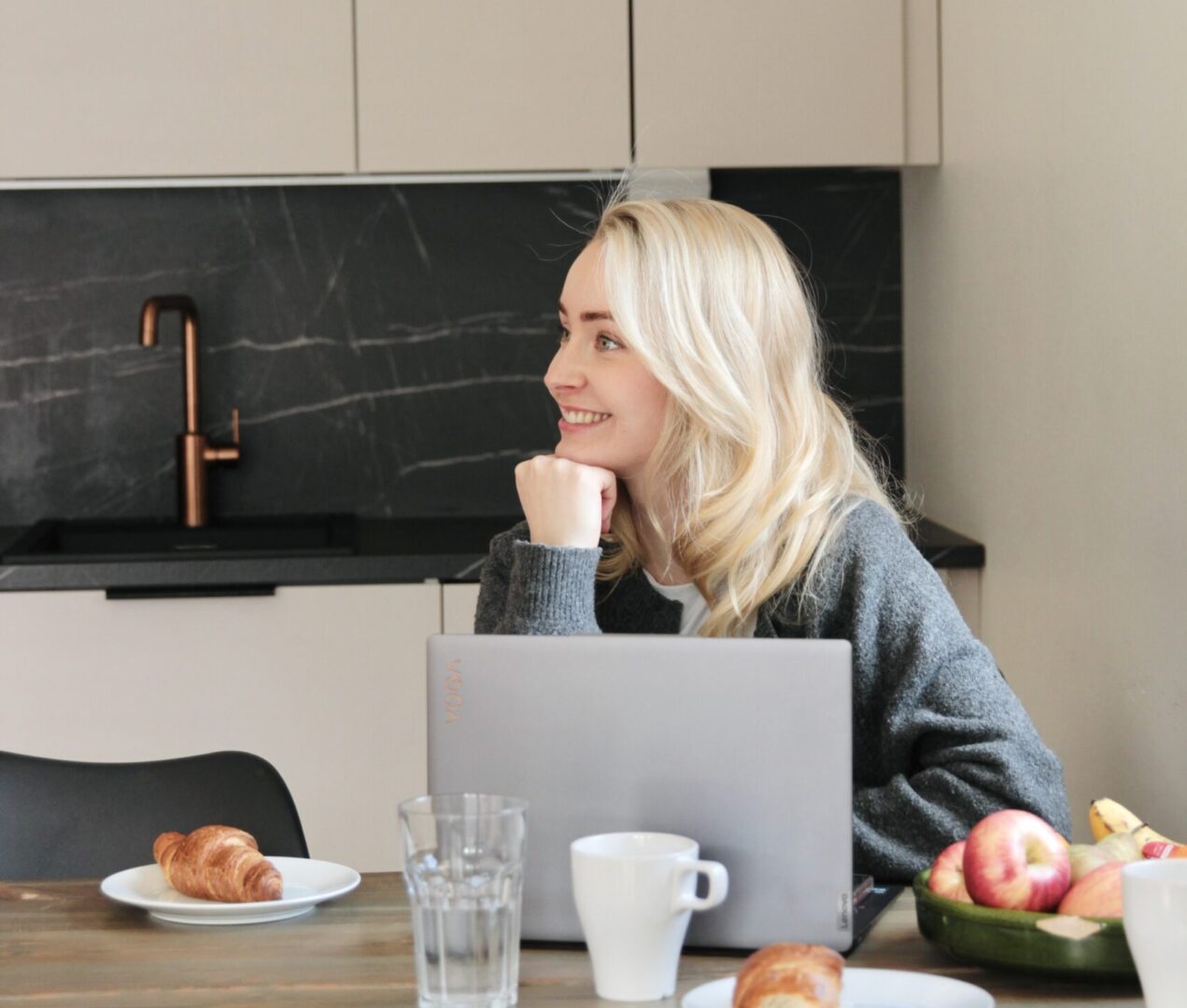 Woman with blonde hair working at a kitchen table with a laptop, croissant on a plate, and a bowl of fruit nearby.