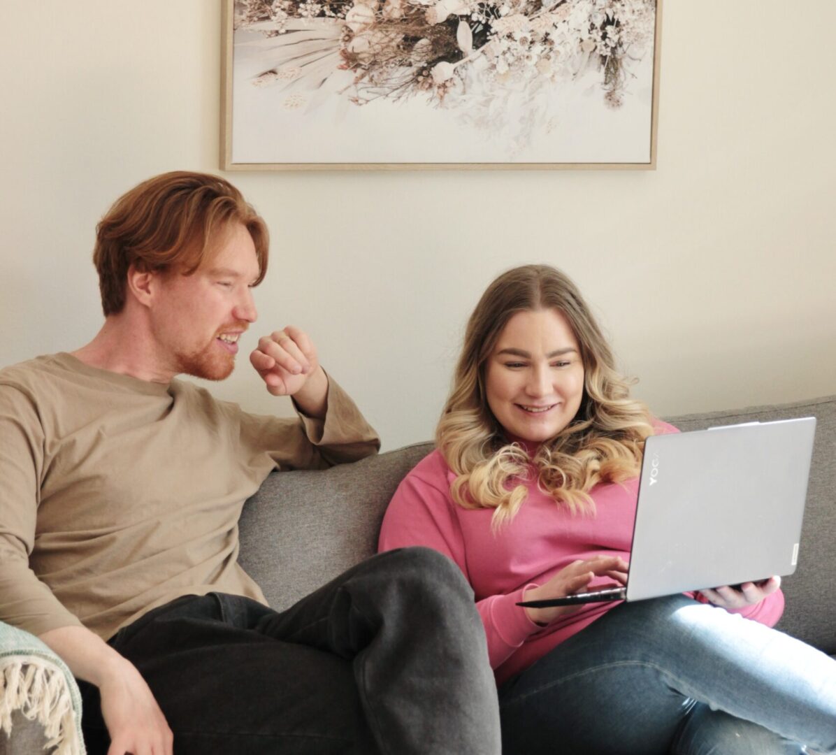 Two adults sit on a gray sofa in a living room; the woman in pink works on a laptop while the man talks beside her art hangs on the wall above.