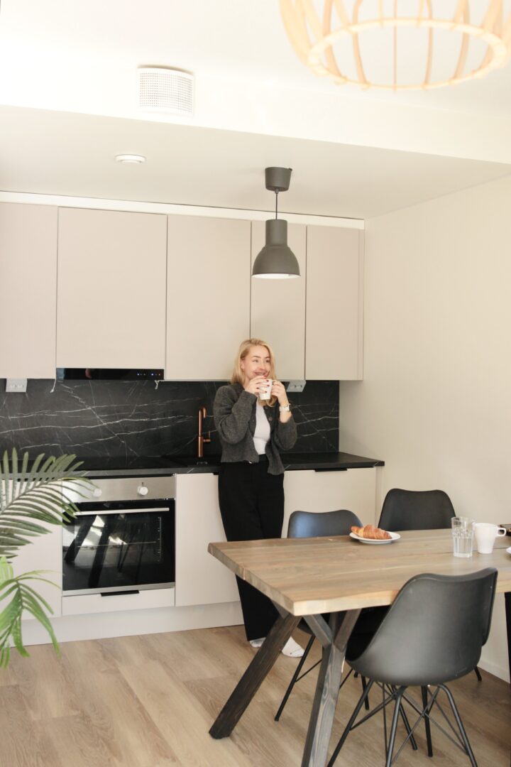 Woman standing in a bright modern kitchen, holding a mug near the counter while a croissant and glasses sit on a dining table in the foreground.