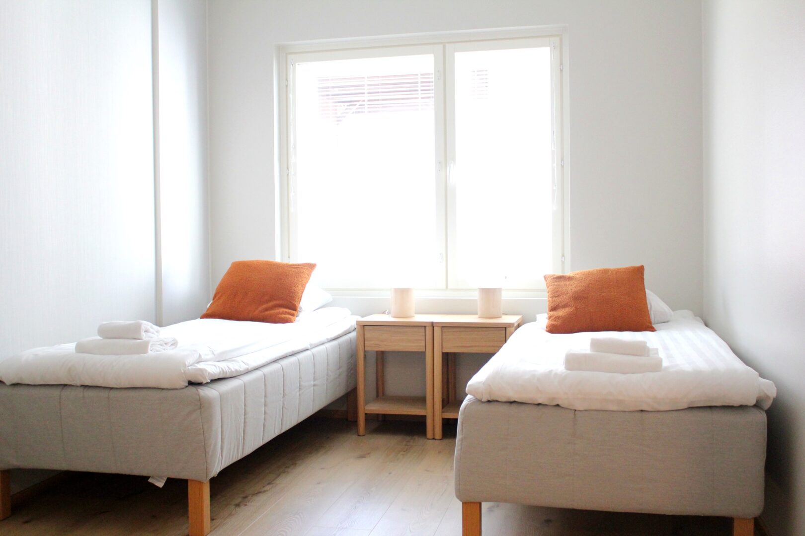 Two single beds with white bedding and orange throw pillows face a central window, a small wooden nightstand between them.