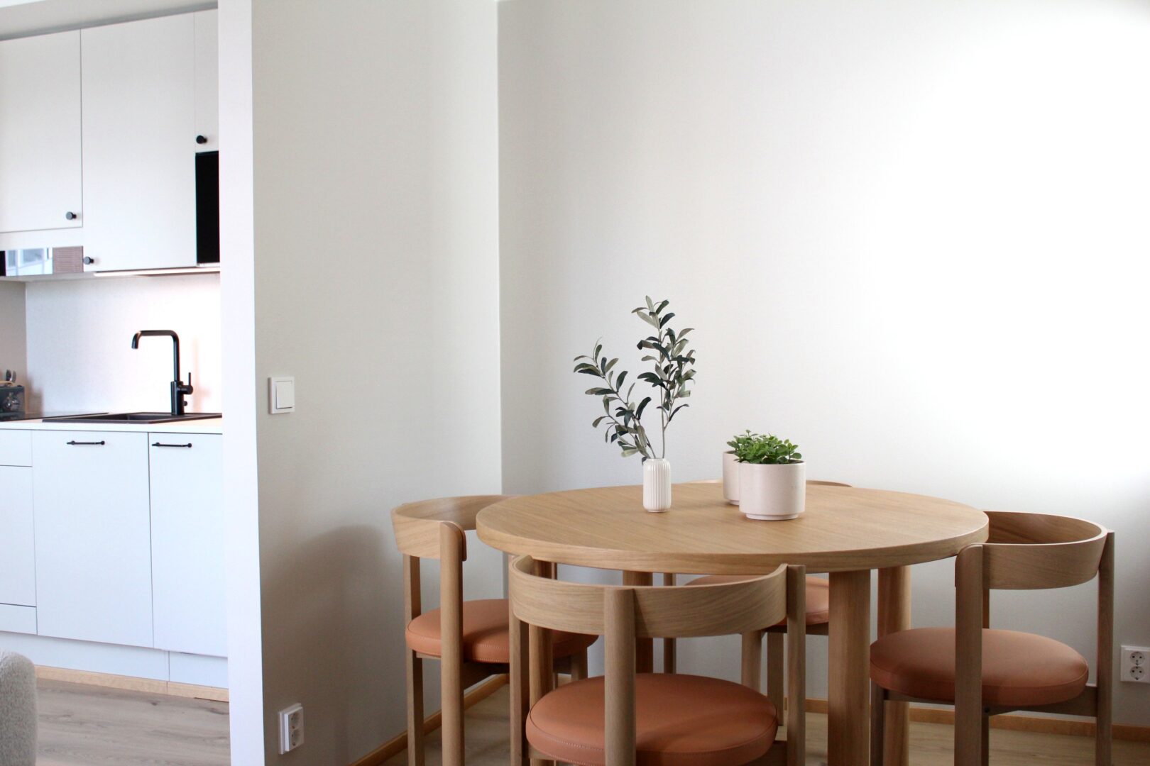 Light-filled kitchen and dining area with white cabinets on the left and a round wooden table with orange-brown stools and potted plants on the table area, against pale walls.