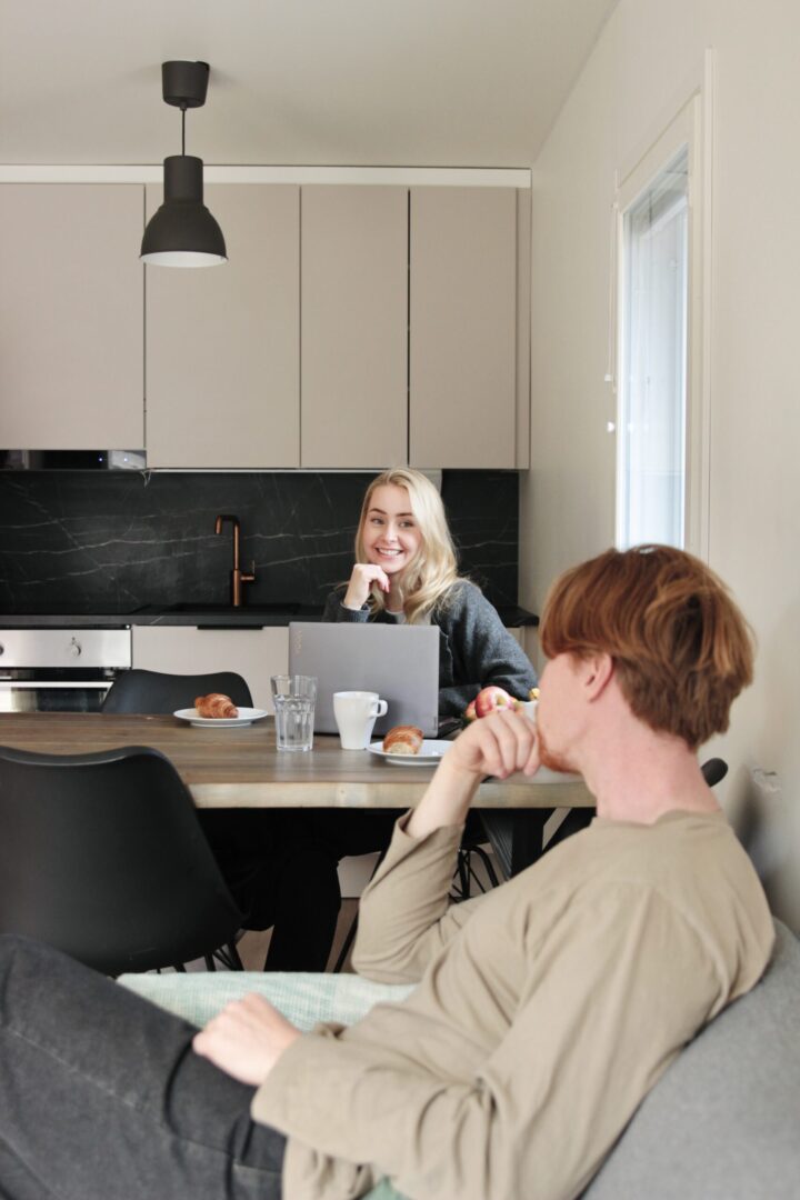 Two people sit at a kitchen island with coffee and pastries; woman smiles at a laptop while the other person sits nearby.