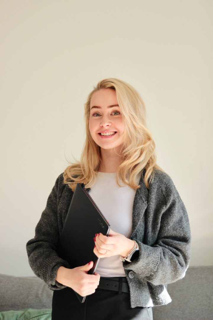 maria-kubier-spot-apartments – Spot Apartments Smiling woman with long blond hair holding a black notebook, wearing a gray cardigan and white top against a light background.