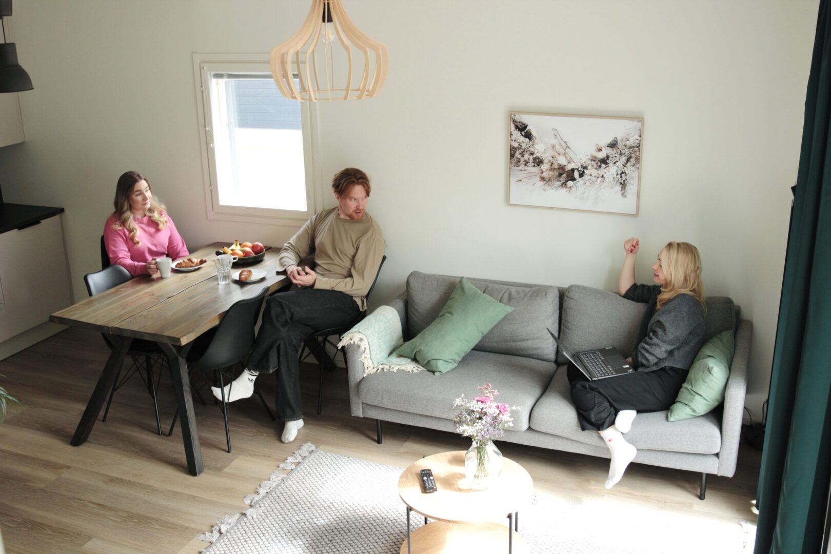 A family breakfast in a bright living room: three people sit around a wooden table with fruit and pastries, sofa on the right, artwork on the wall, window behind.
