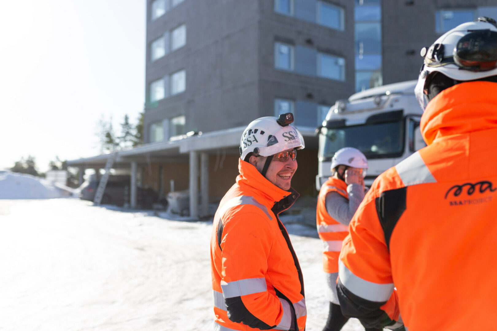 SSA rakennusliikeen rakennustyöntekijät oransseissa huomiovaatteissa ja suojakypärissä keskustelevat kerrostalon edustalla talvisessa ympäristössä. Etualalla oleva työntekijä hymyilee, taustalla näkyy työmaa-ajoneuvoja ja lunta.